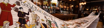 A close up view of a brewery inspired version of the Bayeux Tapestry being unrolled on a long table in a pub.