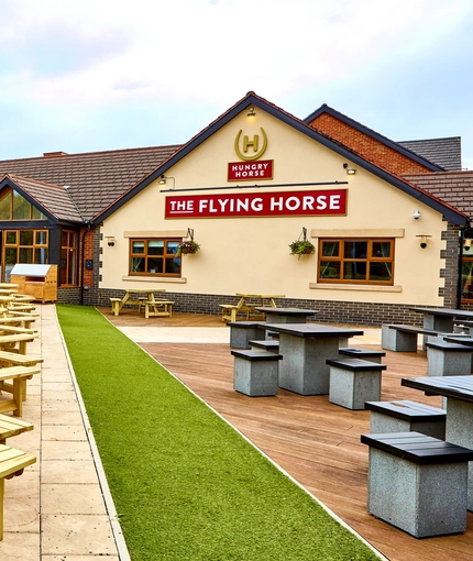 The exterior facade, signage and beer garden seating area of The Flying Horse in Heald Green, featuring wooden picnic tables and a children's play area.