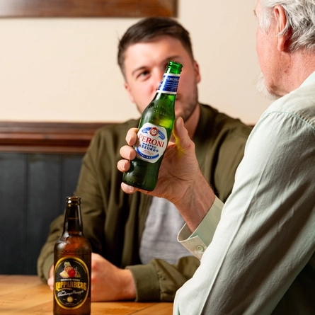 Three people seated at an indoor table. One man is holding a bottle of Peroni, whilst his two companions have their drinks on the table in front of them..  