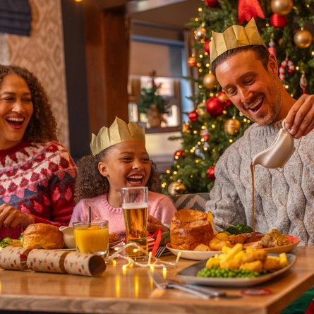 A festive Christmas meal taking place beside a decorated Christmas tree adorned with red and gold baubles, candy cane ornaments and warm fairy lights. Three people seated at a dining table wear paper crowns from Christmas crackers while enjoying a traditional festive meal. One person pours gravy from a gravy boat onto a dinner plate. The table is set with roast dinners, vegetables, glasses of orange juice, a pint of beer, Christmas crackers and string lights, creating a warm and seasonal restaurant setting.