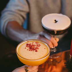 A close up view of two cocktails served in coupe glasses being held by two people sitting at a table in a pub.