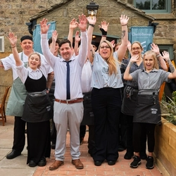 A group of people stood outside a pub, arms raised and smiling at the camera.