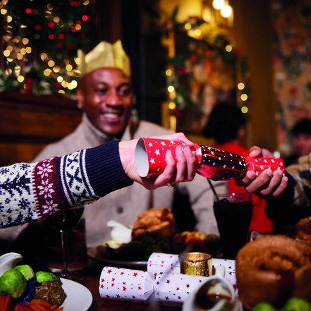 Two hands pulling a Christmas cracker over a table, with a blurred out face of a man looking on and smiling.