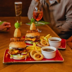 Two stacked burgers topped with festive garnishes are served with chips, onion rings and gravy on red plates at a wooden table. Glasses of white wine, rosé and beer are arranged around the table, with diners seated nearby. The cosy pub setting features warm lighting and festive decorations in the background.