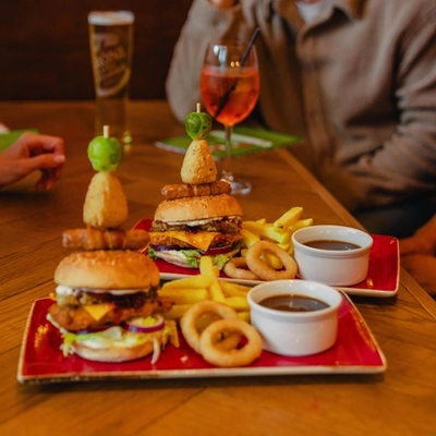 Two stacked burgers topped with festive garnishes are served with chips, onion rings and gravy on red plates at a wooden table. Glasses of white wine, rosé and beer are arranged around the table, with diners seated nearby. The cosy pub setting features warm lighting and festive decorations in the background.