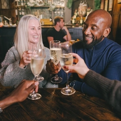A lifestyle image focusing on 2 customers sat at a table with friends and glasses of sparkling white wine, celebrating with a cheers for Mothers Day within the interior seating area at a Heritage venue.