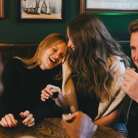 A group of people at a wooden table inside a pub, laughing together and holding shots of Guinness.