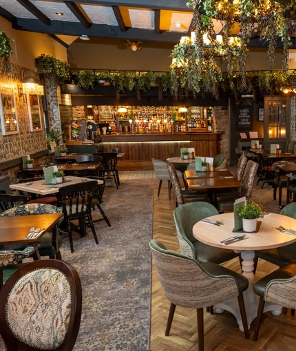 Some tables in The Rockingham Arms restaurant area, with foliage hanging from the windows and lights and the bar in the background.