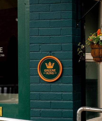 A close up view of the Greene King logo on a glass door of the Malvern Tavern, and on a circular green and yellow plaque on the brick wall next to it.