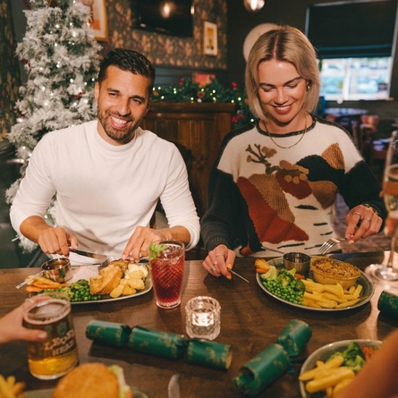 Four people sitting at a restaurant table with plates of food in front of them. Glasses of Level Head IPA, Guinness and a cocktail also sit on the table and one person is raising a glass of champagne.