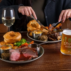 A woman using cutlery to dig into a plate of roast dinner sitting on the table in front of her next to drinks and another roast dinner.
