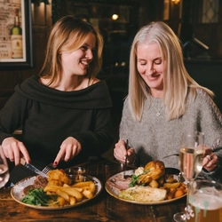 A lifestyle image focusing on 2 customers within a group sat at a table enjoying Mothers Day main dishes and drinks within the interior restaurant seating area at a Heritage venue.