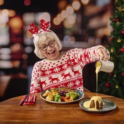 A person wearing reindeer antlers and a Christmas jumper, sitting at a restaurant table and pouring custard over the plate of pudding in front of them. Cutlery, a red serviette, and a plate of Christmas dinner also sit on the table; and a Christmas tree is visible in the background.