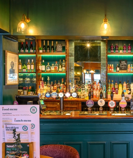 A close up view of one of the wood panelled bars inside The Globe; with golden drinks trays on the counter, and bottles of wine and alcoholic spirits on illuminated shelves behind the bar.
