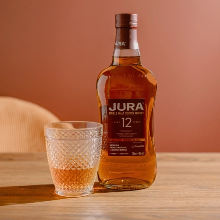 A full bottle of Jura single malt Scotch whisky, standing on a wooden table next to a lowball glass containing some of the whisky.
