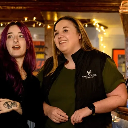 Two Women stood by the bar, the woman on the left is pointing with her arm extended. 