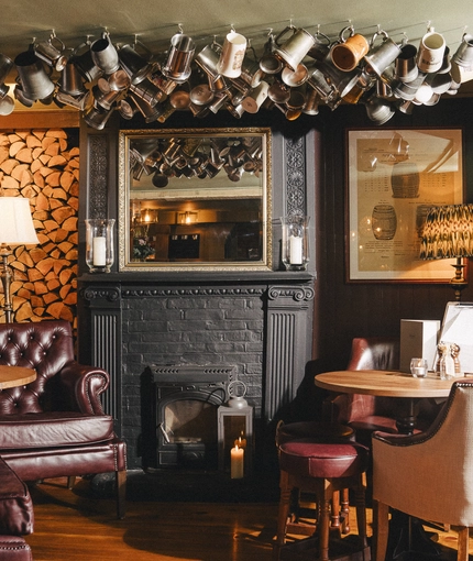 An interior restaurant seating area at Ye Olde Swan in Thames Ditton, with armchairs, a fireplace, and a variety of tankards hanging from the ceiling.