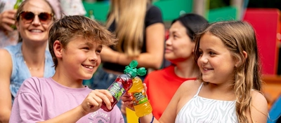Two children sitting next to each other with a bottle of Fruitshoot in a "Cheers" pose.
