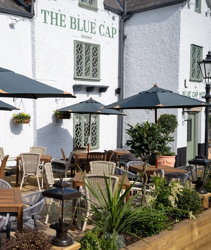 The exterior facade and beer garden seating area at The Blue Cap in Sandiway, with flower baskets hanging on the wall, planter boxes, and shade umbrellas.