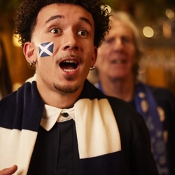 Inside a warmly lit Greene King pub during a Six Nations match day, a person stands in the foreground with a Scottish flag painted on his cheek and a thick navy‑and‑white striped rugby scarf draped around the neck. The individual is surrounded by other supporters, who are gathered closely together in the lively atmosphere typical of match screenings. The background shows warm golden lighting, hanging glassware, and the characteristic décor of a busy pub. The scene captures the sense of anticipation and communal spirit associated with fans watching the Six Nations tournament together.