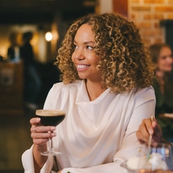 Two people sitting at a restaurant table with a pudding served on a plate, ice cream served in a sundae glass, and a glass of wine in front of them. One person is holding a cocktail and the other person is using a spoon to scoop up some of the ice cream.