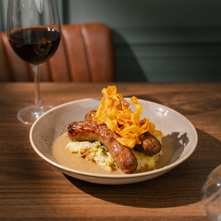 An advertising image featuring a Game Venison Sausage With Apple & Squeak dish sat on a table with a glass of red wine from the Game Festive Menu at Chef & Brewer venues.