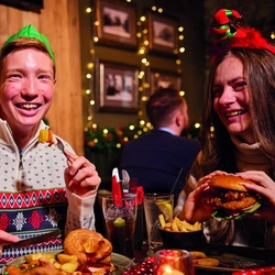 A group of people are gathered around a table enjoying a festive meal. They are wearing paper crowns, and the table is set with plates of food. The background is decorated with Christmas decorations.