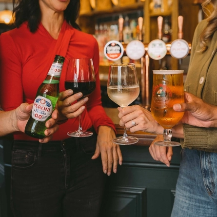 Four people holding drinks at a pub bar, including Peroni 0.0%, red and white wine, and a pint of Birra Moretti lager.