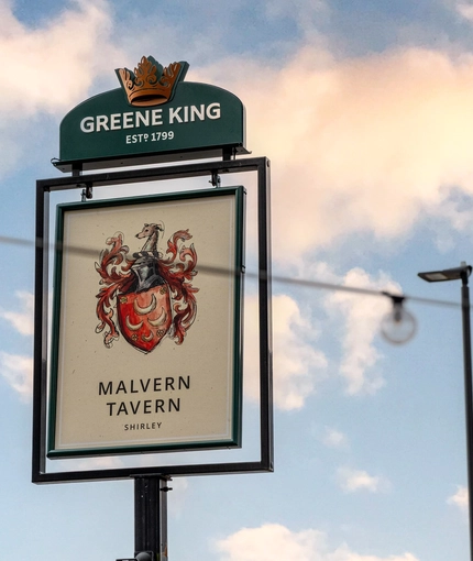 A close up view of the exterior pub sign of the Malvern Tavern, featuring an illustration of a red coat of arms with four crescent moons, filigree patterns and a dogs head, above the words "Malvern Tavern, Shirley".