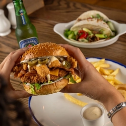 Two hands holding a Crispy Double Chicken Ranch Burger over a plate on a wooden table.