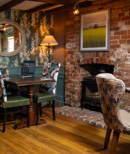 An interior restaurant seating area at The Kings Arms in Sandford-On-Thames, with wooden flooring and beams, upholstered chairs, and a brick fireplace with a log burner.