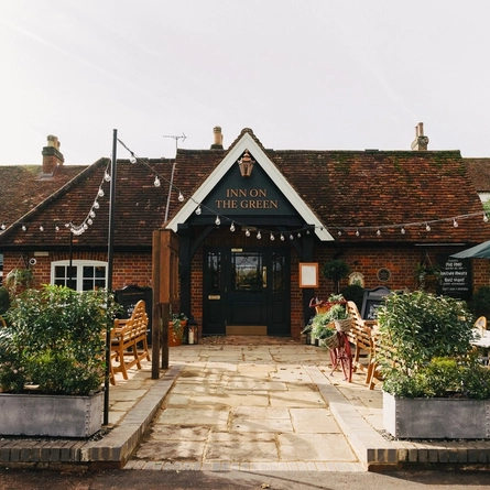 The exterior facade, signage, and beer garden seating area of The Inn On The Green in Harpenden, with wooden furniture, shade umbrellas, shrubs in planter boxes, and string lights overhead.