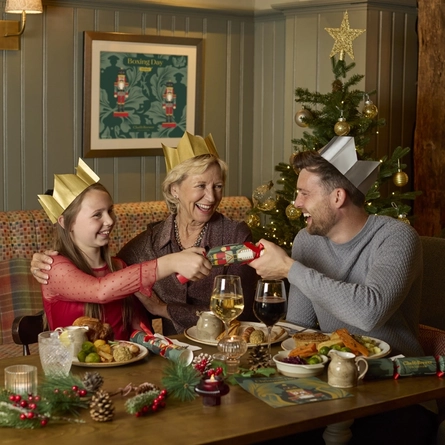 An image showing 2 adults and a child sat within the interior restaurant seating area pulling a cracker while enjoying Christmas Day dishes at a Chef & Brewer venue.