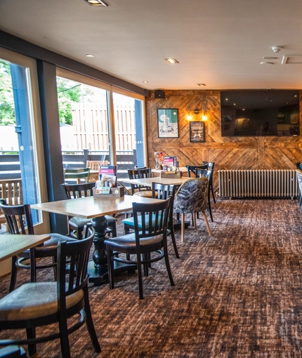 The interior restaurant seating area of The Merlin, with upholstered booth seating, wooden tables, framed artwork and a TV on the walls, and a radiator.