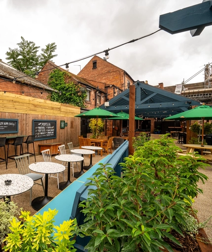 The exterior beer garden seating area at The Talbot in Worcester; with wood picnic tables, shade umbrellas, planter boxes, string lights overhead, and a large wooden pergola with under cover seating.