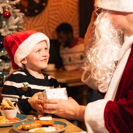 A child wearing a Santa hat sits at a table in a pub decorated for Christmas, receiving a wrapped gift from a person dressed as Santa Claus. An adult sits beside the child, while a snow-covered Christmas tree with red baubles stands in the background. Breakfast plates with milk, fruit and cooked food are visible on the table, creating a festive family dining scene.
