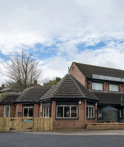 The exterior facade, signage, and seating area at the Devonshire Arms in Sheffield, with an accessibility ramp, wooden picnic tables, shade umbrellas, and string lights.