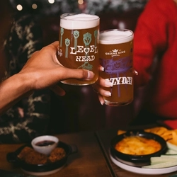 A close up view of two people clinking together glasses of Level Head and Hazy Day, above a wooden restaurant table laden with Christmas crackers and plates of food. A Christmas garland lit with fairy lights is visible in the background.