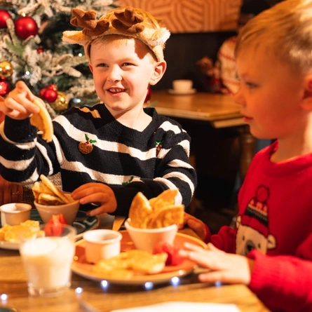 An adult wearing a Santa hat sits with two children at a wooden table in a pub decorated for Christmas. The table is set with glasses of milk, pancakes, strawberries and bowls of crisps, while a snow-covered Christmas tree with red baubles stands nearby. A festive Santa figure and other guests are visible in the background, creating a warm family Christmas breakfast setting.