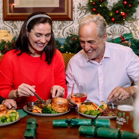 Three people seated around a table in a festively decorated pub, enjoying a traditional Christmas meal. Plates containing roast dinners with Yorkshire puddings, vegetables and roast potatoes are served alongside a pint of beer, glasses of red and rosé wine, and green Christmas crackers. Seasonal garlands, framed artwork, patterned wallpaper and warm decorative lighting create a festive dining atmosphere.