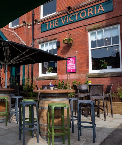 The exterior beer garden seating area at the front of The Victoria, with bar stools, shade umbrellas, planter boxes under the windows, and tables made from large barrels.