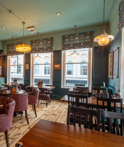 The interior restaurant seating area at The Plough, with upholstered chairs and booth seats, wooden floor, and framed artwork on the wood panelled wall.