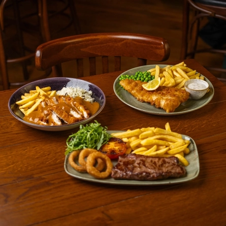 A close up view of a wooden restaurant table laden with a bowl of Katsu Chicken with fries and rice, a plate of Fish & Chips served with peas, lemon, and dipping sauce, and a plate of steak, chips, onions rings, tomato, and salad.