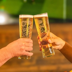 Two hands clink glasses of Peroni Nastro Azzurro lager in a celebratory toast. The chilled beers are served in branded pint glasses with foamy heads, set against a softly blurred pub background in warm green and golden tones.