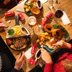 Overhead view of a festive dining table set with Christmas meals and drinks in a pub. Plates include a traditional roast dinner with turkey, Yorkshire puddings, roast potatoes, vegetables and gravy, alongside a steak meal with chips, peas and grilled tomato. Glasses of lager and red wine, Christmas crackers and shared tableware complete the festive dining scene.