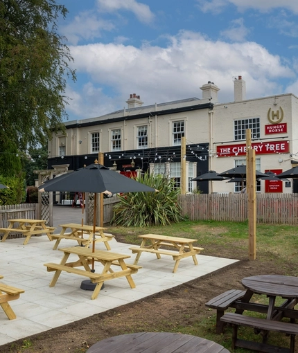 The exterior facade, signage, and beer garden seating area at The Cherry Tree in Newcastle-Under-Lyme; with wooden picnic tables, shade umbrellas, and string lights overhead.