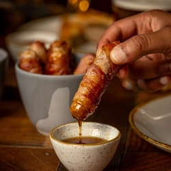 A wooden table with a small bowl of Pigs in Blankets.  A hand is holding a ping in blanket over a small pot of dipping sauce.  