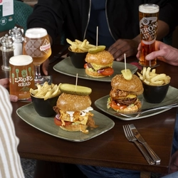 A table with three plates of burgers topped with pickles and served with fries in small cups. Several drinks—including pints of beer—are placed around the table, and the hands and arms of multiple people seated together are visible near the food.