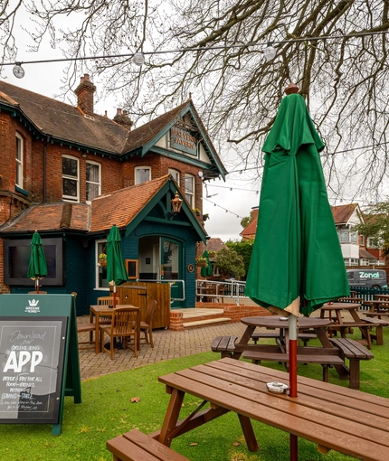 The exterior facade, signage, and beer garden seating area of the Malvern Tavern in Shirley, with wooden picnic tables, shade umbrellas, string lights, and an accessibility ramp to the entrance.