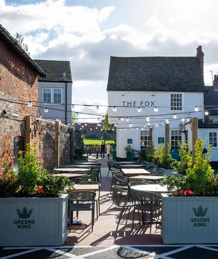 The exterior facade and beer garden seating area of The Fox in Bury St Edmunds, with plants in raised planter boxes and string lights above the tables and chairs.